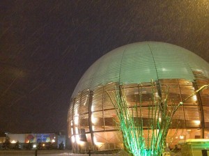 The CERN Globe, partially covered in snow, as more snow falls. The ATLAS Control Room building is seen in the distance.