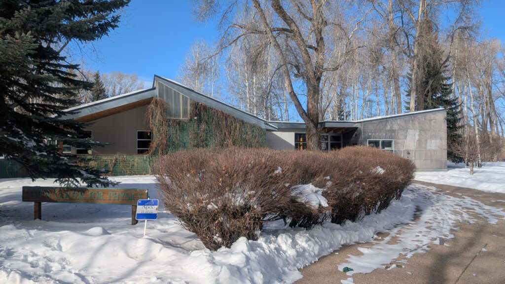 A building at the Aspen Center for Physics campus. Snow is on the ground, the sky is blue, and the building has a modern stone alpine feel to it.