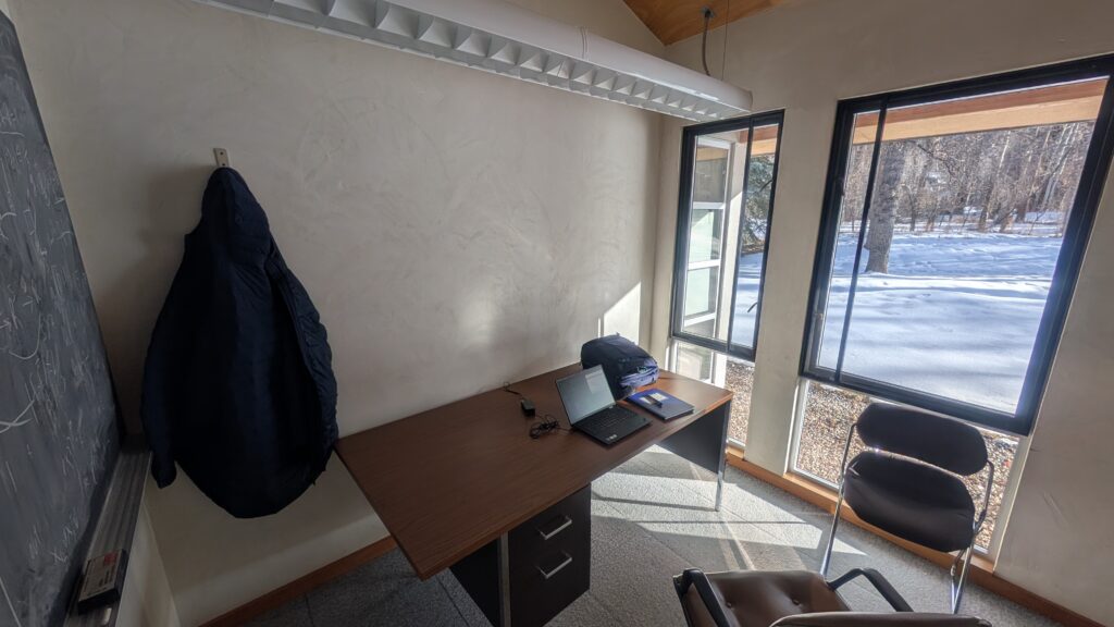 A general-use office at the Aspen Center for Physics. There is a blackboard, a desk and chairs, and a window looking out onto a snowy lawn with trees.