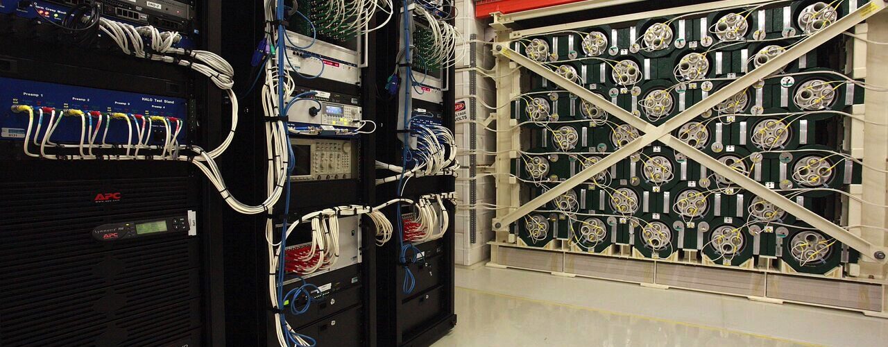 Racks of electronics stand next to and just in front of a large, cube-shaped experiment. The experiment is made from green-painted lead and 128 tubes filled with helium-3.