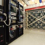 Racks of electronics stand next to and just in front of a large, cube-shaped experiment. The experiment is made from green-painted lead and 128 tubes filled with helium-3.