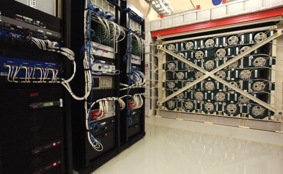 Racks of electronics stand next to and just in front of a large, cube-shaped experiment. The experiment is made from green-painted lead and 128 tubes filled with helium-3.